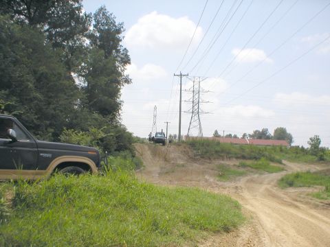 1983 Ford Bronco - A dark brown and tan Ford Bronco is partially visible on the left, with another vehicle in the distance on a dirt track.