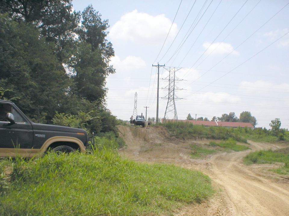 Dark Ford Bronco parked on grass beside a dirt trail with power lines and a pickup in the background