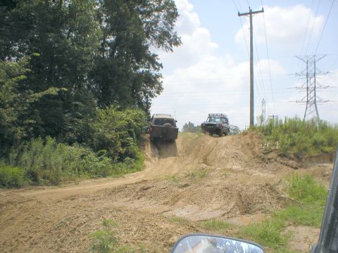 1983 Ford Bronco - Two off-road vehicles navigate a challenging muddy incline under a partly cloudy sky.