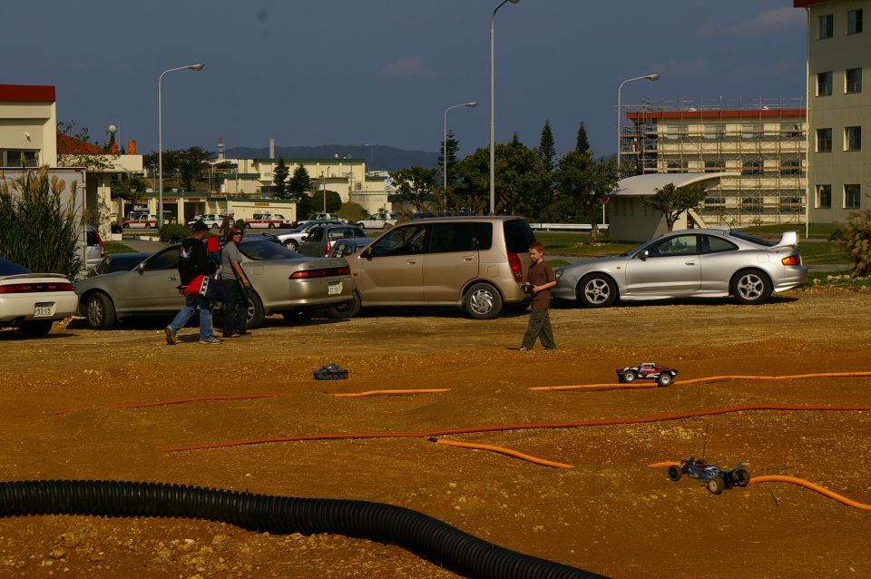 Remote control cars race on a dirt track with parked cars in the background