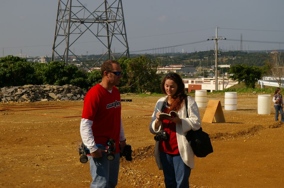 Man in red shirt holding RC car, woman in white cardigan writing in notebook. Outdoor dirt area with power lines and buildings in background