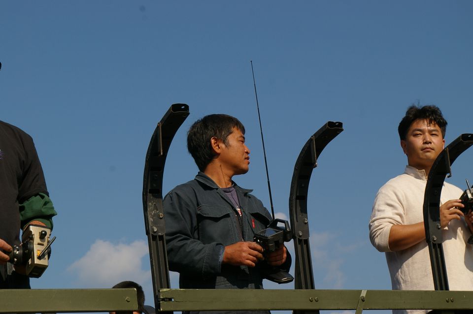 Three men stand outdoors holding remote control devices with antennas