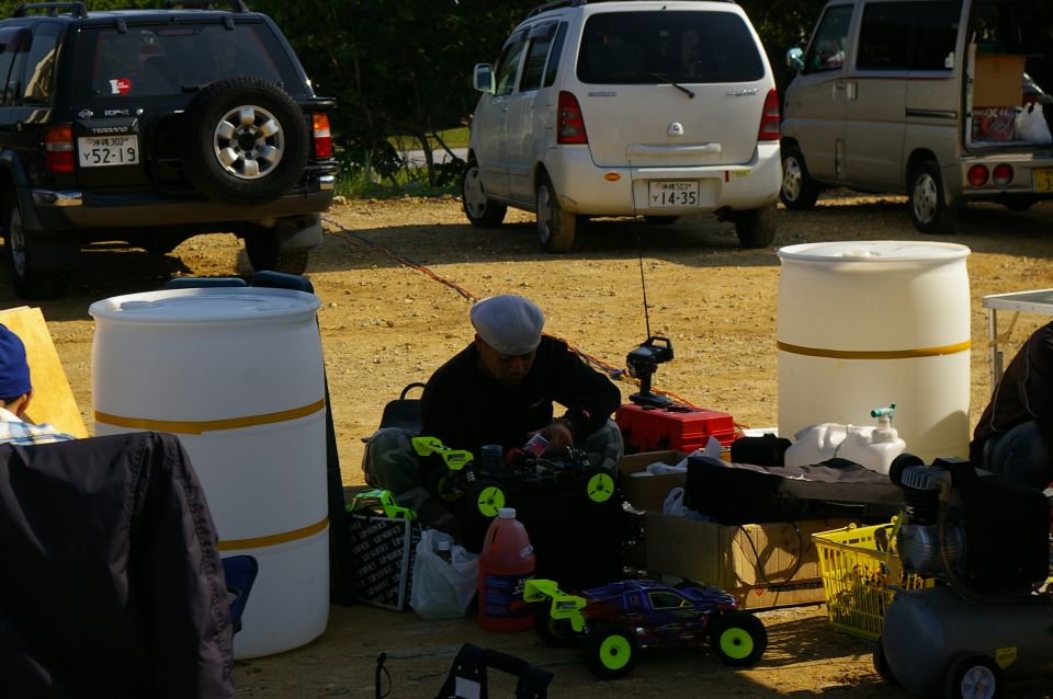 Black SUV with spare tire on rear, white minivan, and silver van parked on dirt lot with people and RC cars