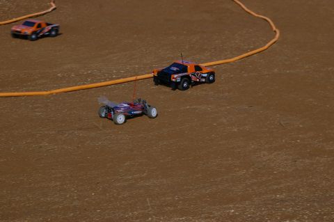 1986 Ford Bronco - Remote control cars compete on a dirt track, with one orange and black buggy in the foreground and another orange truck in the background.