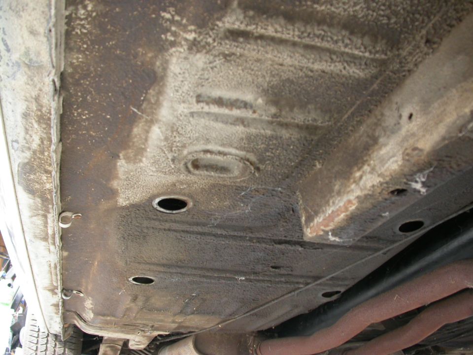 Underside of a car showing the floor pan, frame rails, and exhaust pipes. Some rust and dirt are visible
