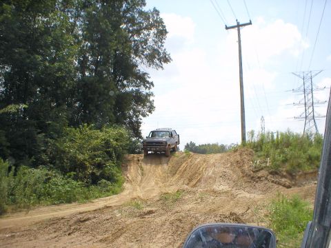 1983 Ford Bronco - A dark SUV navigates an off-road course, climbing a steep dirt ramp under a cloudy sky.
