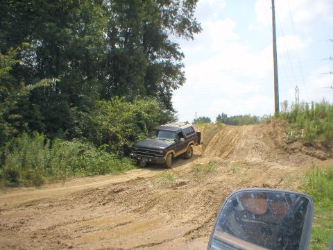 1983 Ford Bronco - A Ford Bronco navigates a muddy incline on a dirt road, surrounded by greenery and under a partly cloudy sky.