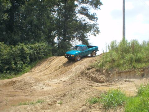 1983 Ford Bronco - A teal pickup truck navigates a challenging dirt incline, kicking up dust and mud.