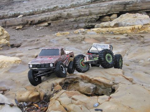 1993 Toyota 4Runner - Two RC rock crawlers are positioned on a rocky coastline, with one truck in the foreground and another behind it.