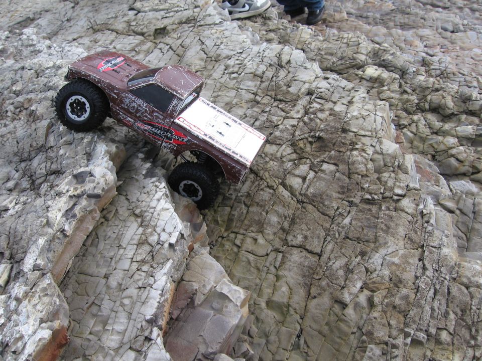 RC rock crawler truck on a rocky slope. Some people's feet are visible at the top