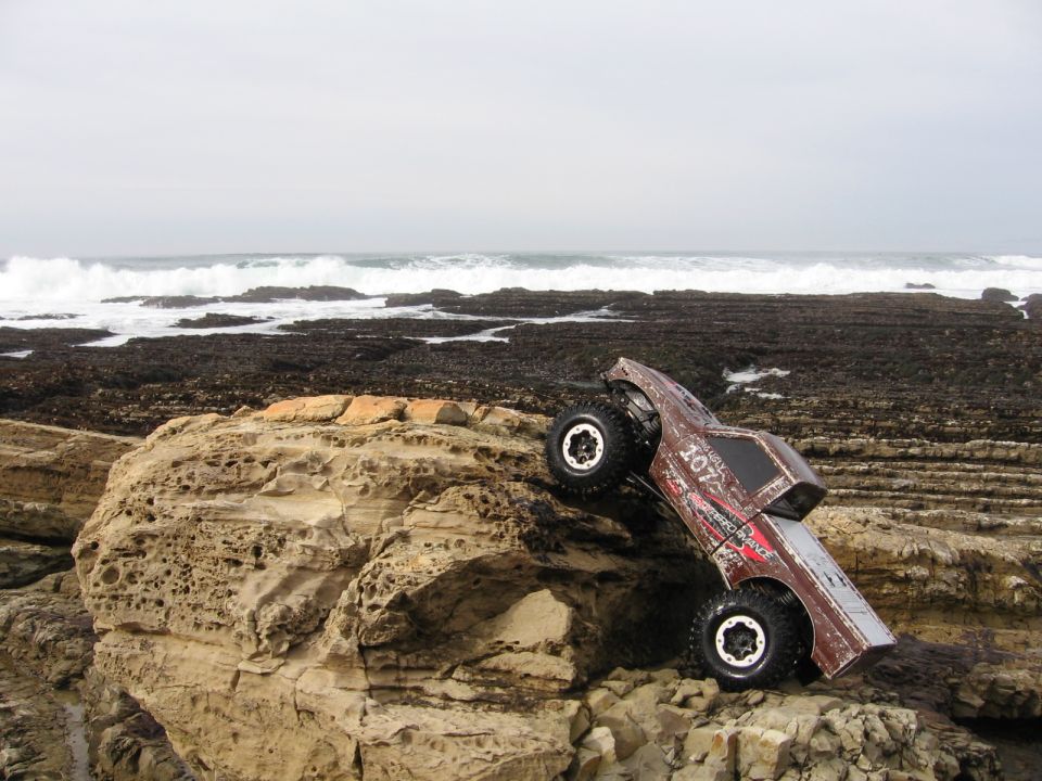 Brown remote control rock crawler with number 107 on its side, climbing rocks near the ocean