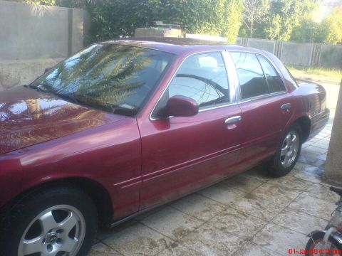 1998 Ford Crown Victoria - A red 1998 Ford Crown Victoria sedan is parked on a tiled surface with trees and a wall in the background.