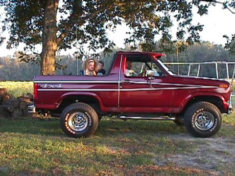 1984 Ford Bronco - Side view of a red SUV with people inside and in the open bed, parked outdoors.