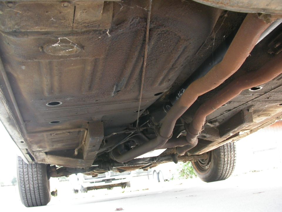 Underside of a rusty car with dual exhaust pipes and a driveshaft. Another vehicle is visible in the background