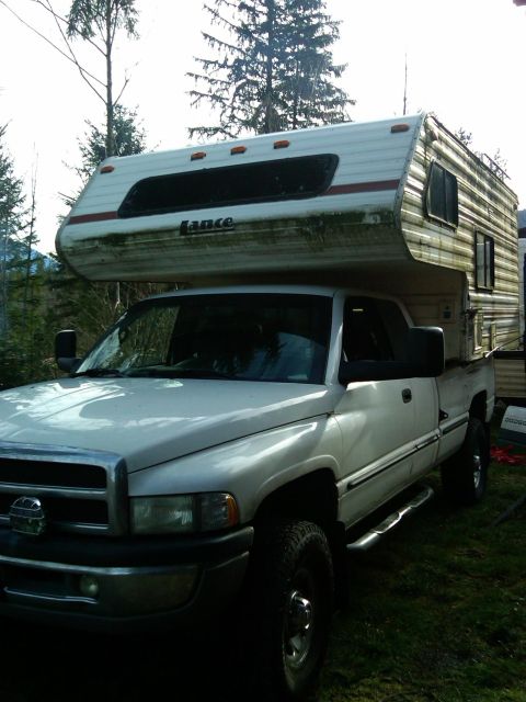 1978 Ford Bronco - A white Dodge Ram pickup truck is fitted with a Lance brand camper shell, parked outdoors.