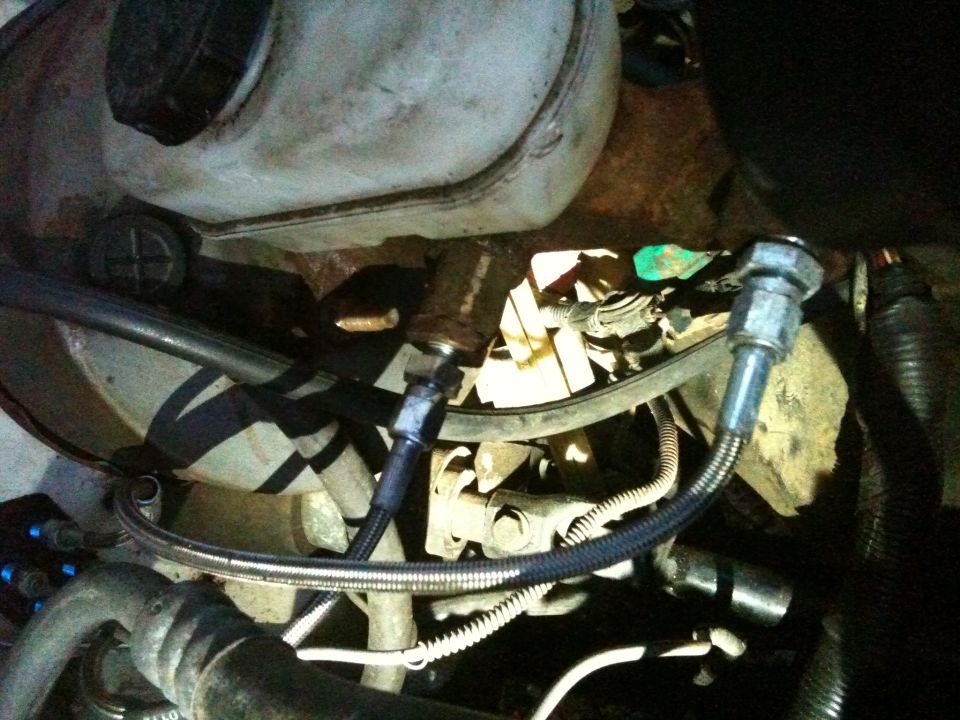 Two braided steel lines with hex fittings in a rusty engine bay near a fluid reservoir