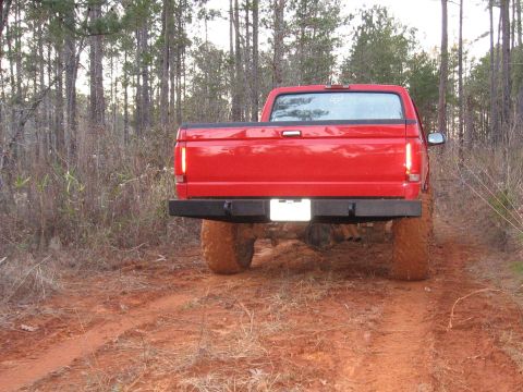 1995 Ford F-150 - A red pickup truck is shown from the rear, driving on a muddy dirt road through a forest.