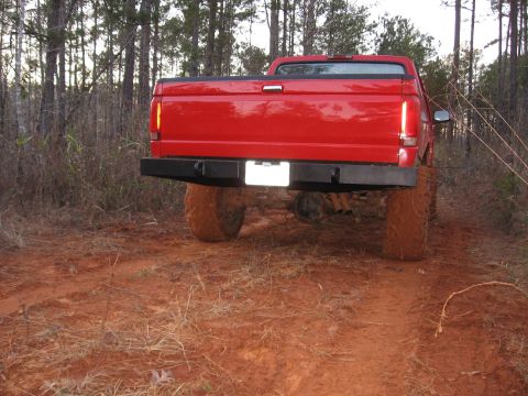 1995 Ford F-150 - Rear view of a lifted red pickup truck with a black bumper and large tires driving on a dirt path through a wooded area.