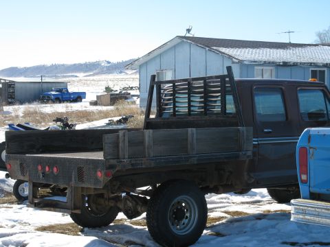 1977 Ford F-250 - A brown flatbed truck with wooden sides is parked in a snowy area next to a building. Another blue truck is visible in the background.
