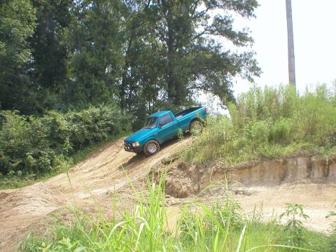 1983 Ford Bronco - A teal pickup truck navigates a muddy, off-road trail surrounded by dense vegetation.