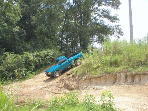 1983 Ford Bronco - A teal pickup truck navigates an off-road incline with trees and brush in the background.