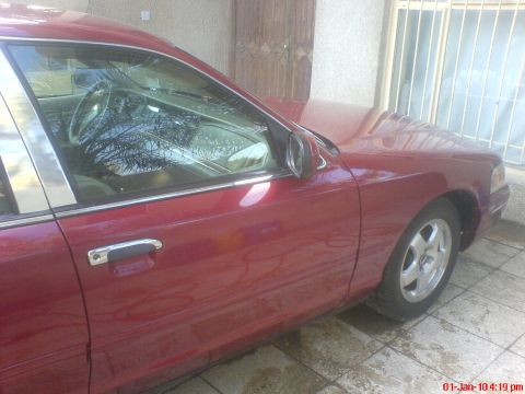 1998 Ford Crown Victoria - A red 1998 Ford Crown Victoria sedan is shown from the side, parked near a building with barred windows.