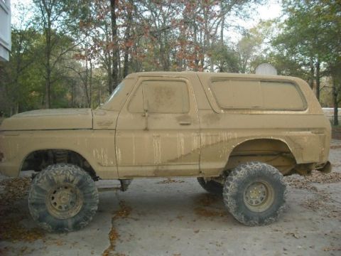 1979 Ford Bronco - A heavily muddied 1978 Ford Bronco sits on a paved surface, its side profile clearly visible with large off-road tires.