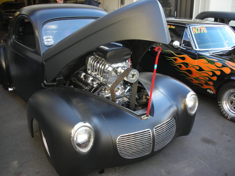 Matte black hot rod coupe with a supercharged V8 engine. Another car with flame graphics is next to it