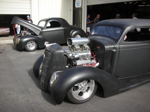 1992 Ford Crown Victoria - Custom hot rods with exposed engines are parked outside a building, with people milling around.