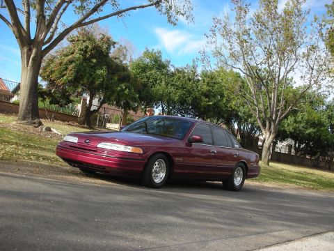 1992 Ford Crown Victoria - A maroon 1992 Ford Crown Victoria is parked on the side of a street with trees and houses in the background.