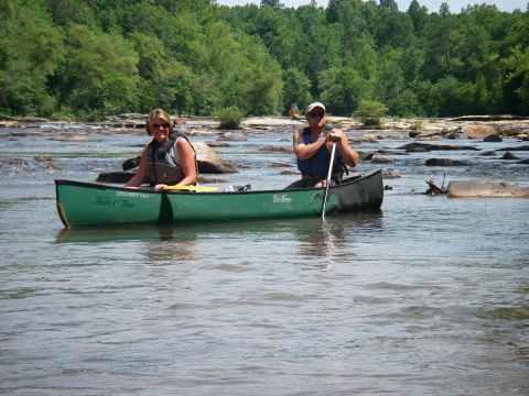 1978 Ford Bronco - Two individuals navigate a river in a green canoe, surrounded by rocky rapids and lush trees.