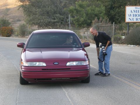 1992 Ford Crown Victoria - Head-on view of a lowered maroon Ford Crown Victoria sedan sitting low on a rural two-lane road, a person visible in the driver's seat wear…