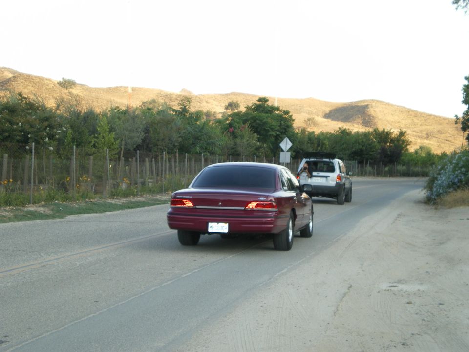 Dark red Ford Crown Victoria sedan from the rear on a rural road with dry hills behind
