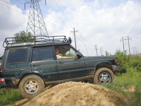 1983 Ford Bronco - A man drives a green Land Rover Discovery off-road, navigating a dirt hill under a cloudy sky with power lines overhead.