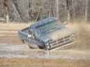 1979 Ford Bronco - A dark-colored pickup truck navigates a muddy terrain, its wheels kicking up dirt as it drives.