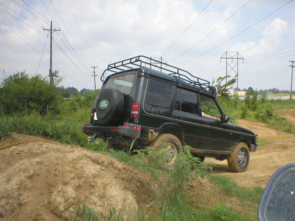 Dark green Land Rover Discovery with roof rack on a muddy off-road dirt trail, rear 3/4 view
