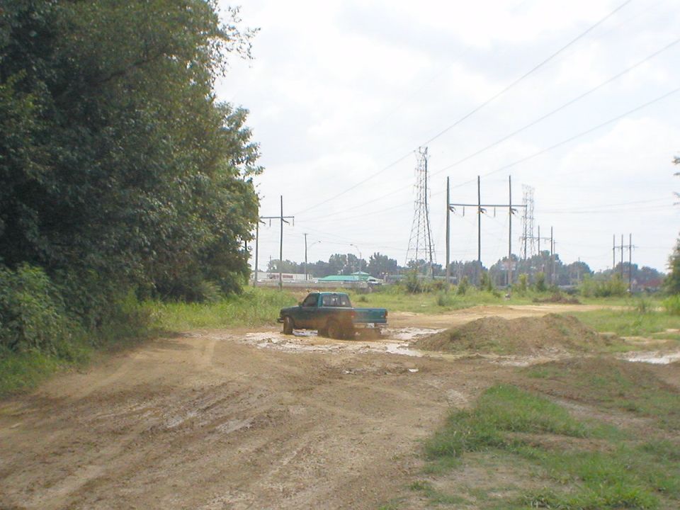 Green pickup truck stuck in mud in a dirt lot with power lines behind.