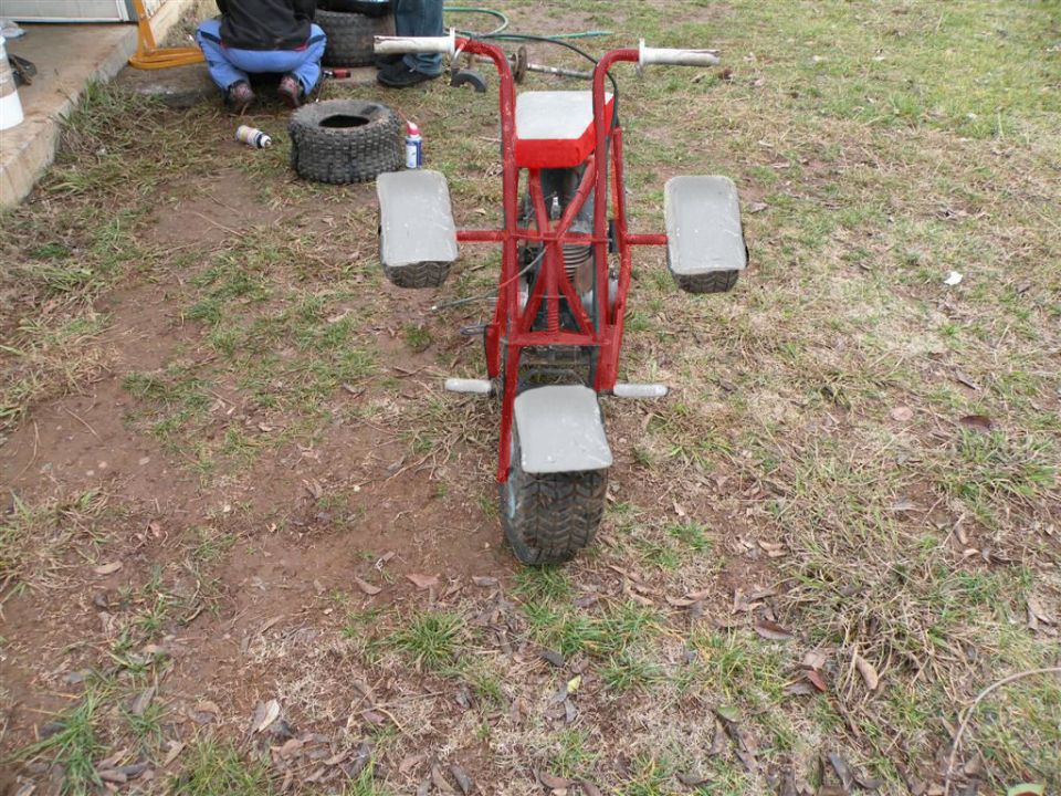 Red mini bike/trike with knobby tires, viewed from rear on grass