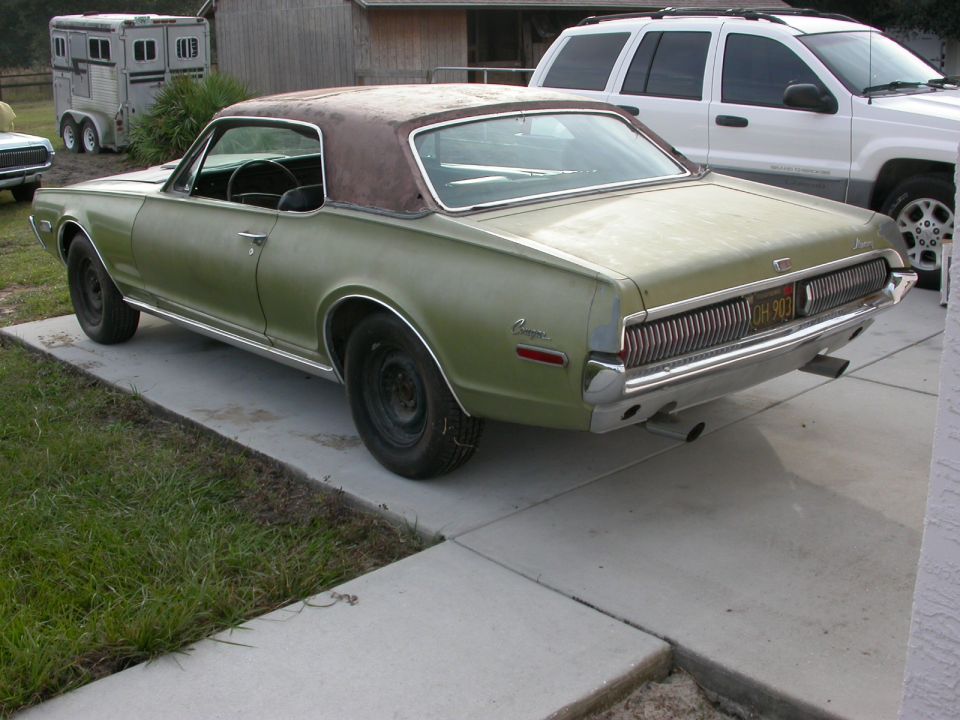 Olive green 1967 Mercury Cougar with a brown vinyl top, parked on a concrete driveway next to a white SUV