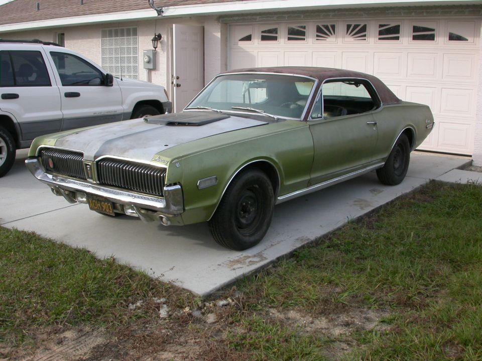 Green 1968 Mercury Cougar with a silver hood and brown roof, parked in a driveway next to a white SUV