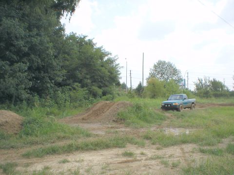 1983 Ford Bronco - A teal pickup truck navigates a dirt track with grassy areas and dirt mounds.