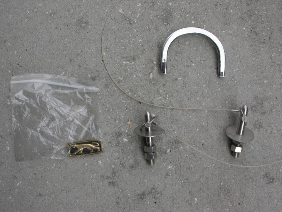 Gold jaguar emblem in plastic bag, chrome U-shaped piece, and two threaded bolts with nuts on concrete