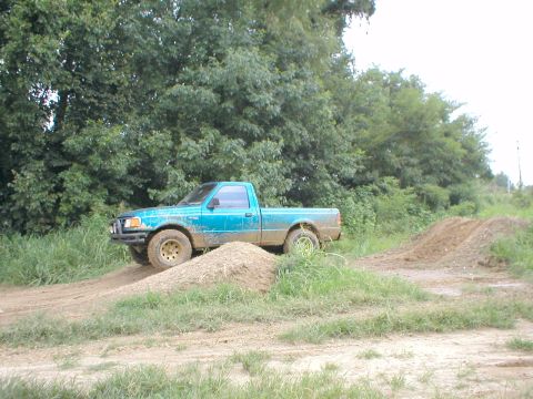 1983 Ford Bronco - A teal pickup truck navigates a dirt track with mounds of dirt.