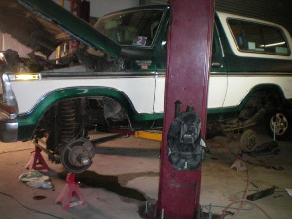 Green and white 1979 Ford Bronco on jack stands in a garage, wheels removed, hood open, front suspension exposed