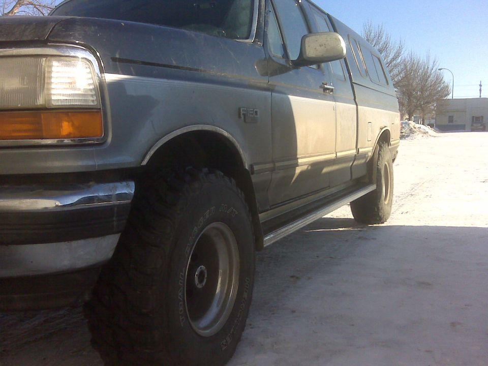 Gray and white Ford F-150 pickup truck with a camper shell, seen from the front quarter angle on a snowy surface