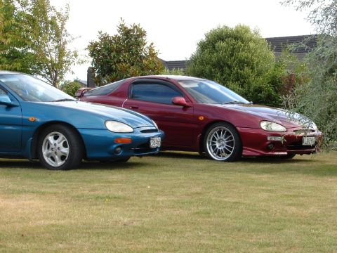 1993 Mazda MX-3 - Two Mazda MX-3s, a blue one and a maroon one, are parked side-by-side on a grassy lawn with trees in the background.