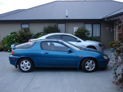 1993 Mazda MX-3 - A blue 1993 Mazda MX-3 coupe is parked next to a silver sedan in front of a house.