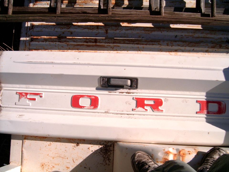 White Ford tailgate with red lettering, showing rust and wear. A person's legs and feet are visible in the lower frame