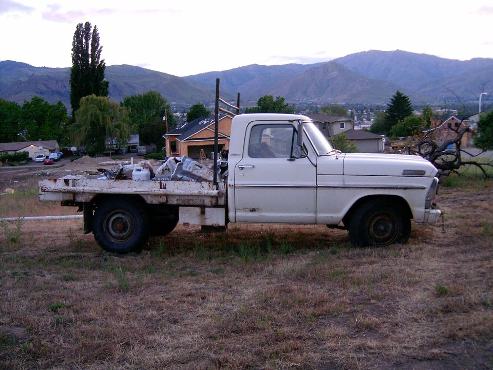 White flatbed truck parked on dry grass with mountains in background. Truck has a ladder rack and is loaded with materials