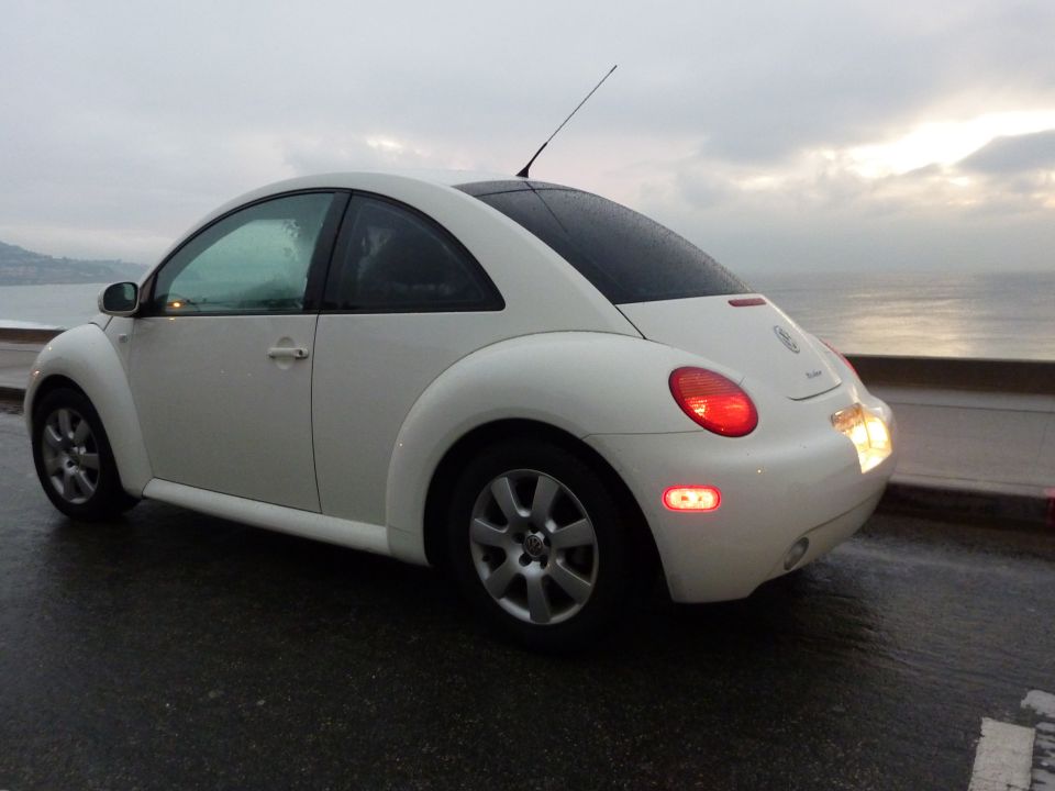 White VW New Beetle rear 3/4 view with brake lights on, parked by ocean at dusk